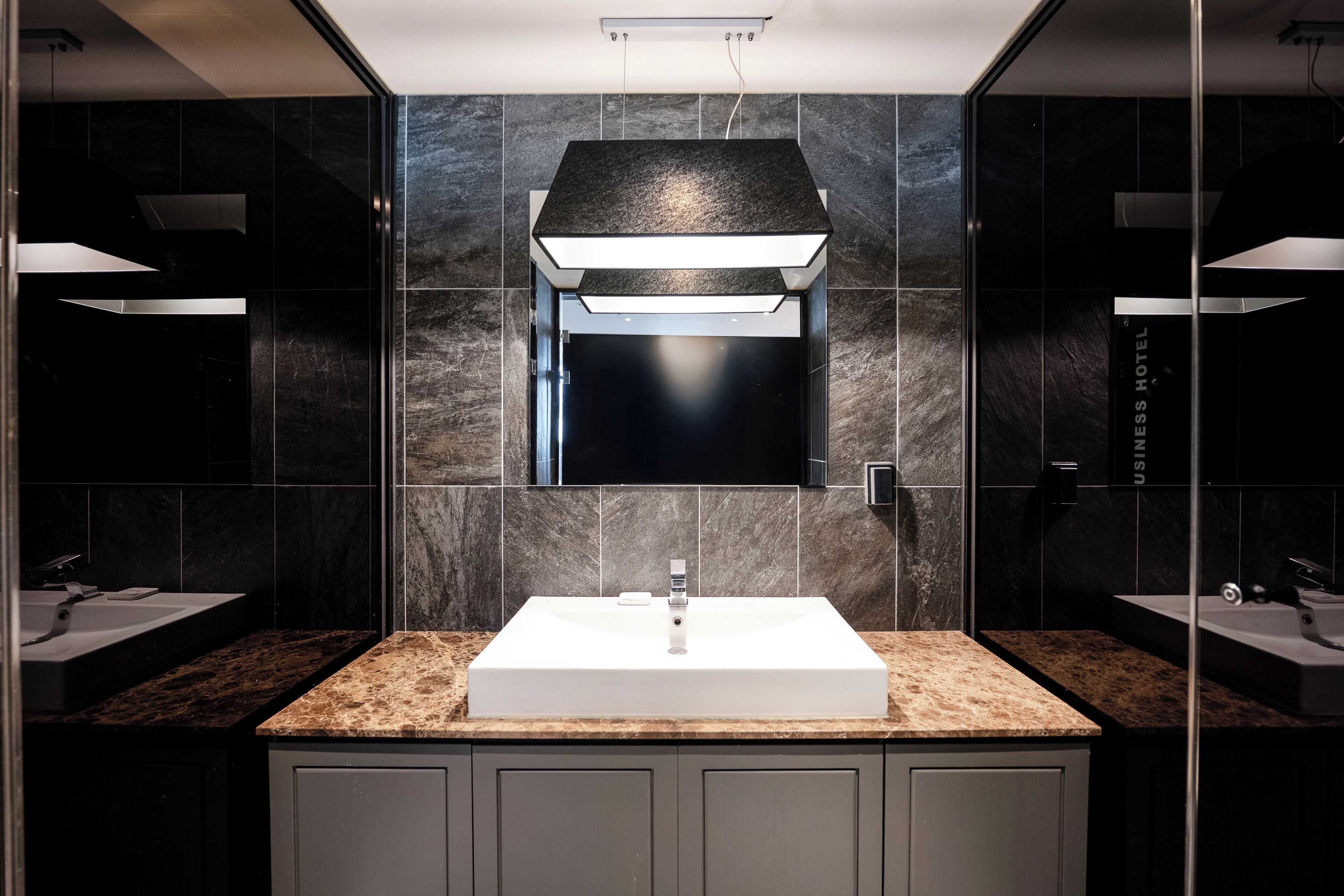 A modern bathroom with a central sink, dark marble tiles, and a large mirror.