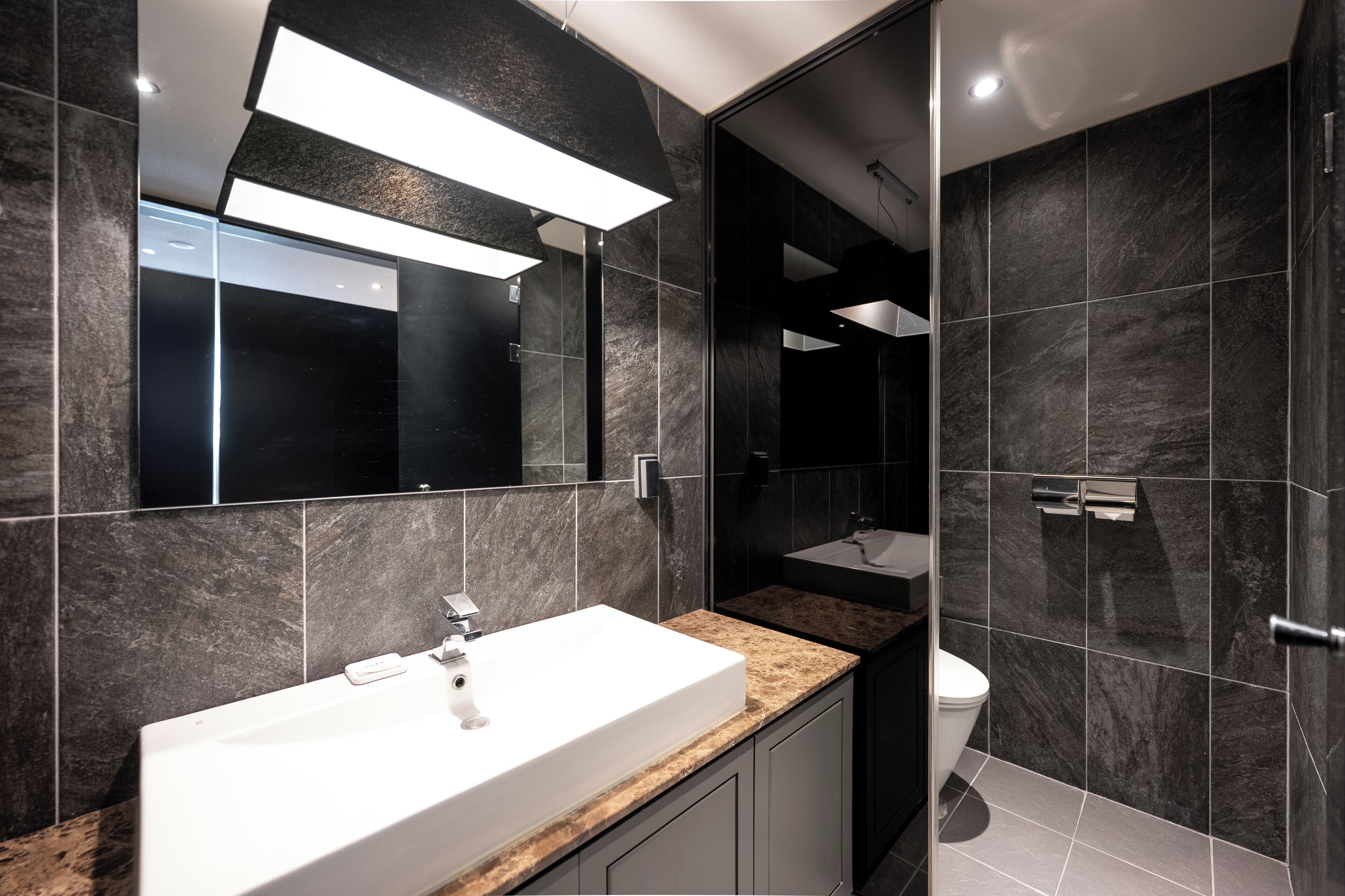 A modern bathroom with a white sink, dark marble tiles, and a large mirror.