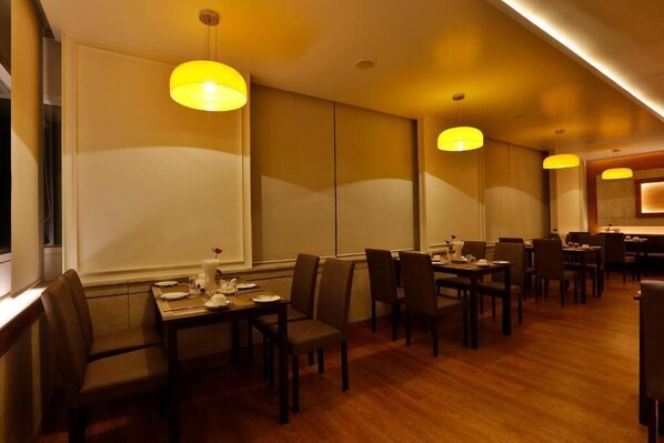 Um restaurante com mesas e cadeiras de madeira, luminárias pendentes amarelas e uma grande janela.