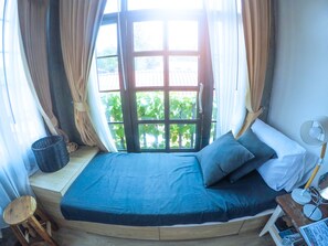 Quarto com cama curva, janela com cortinas, mesa pequena com abajur e cesta.