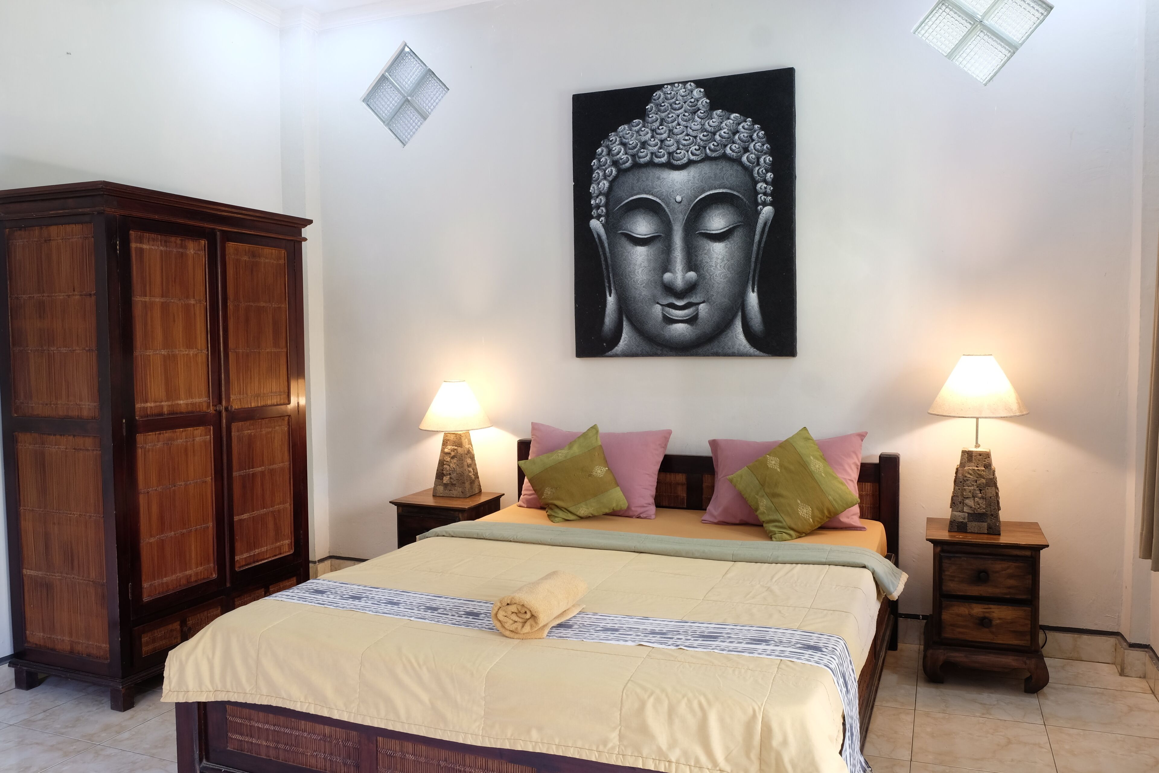 A bedroom with a bed, two nightstands with lamps, a large Buddha painting on the wall, and a wooden wardrobe.