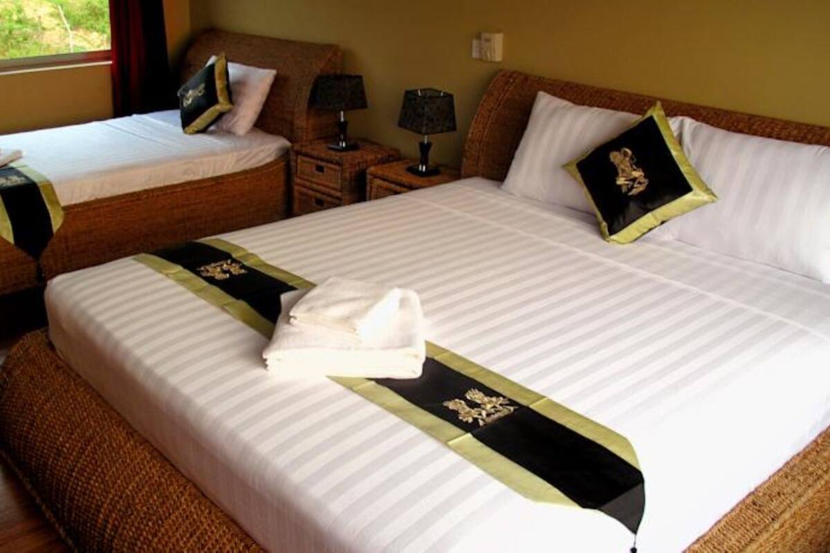 Een hotelkamer met twee bedden, elk voorzien van een witte spreidek en zwarte en gouden accenten. Er staan twee nachtkastjes met lampen, een raam met uitzicht op groen, en een bruine geweven bank.