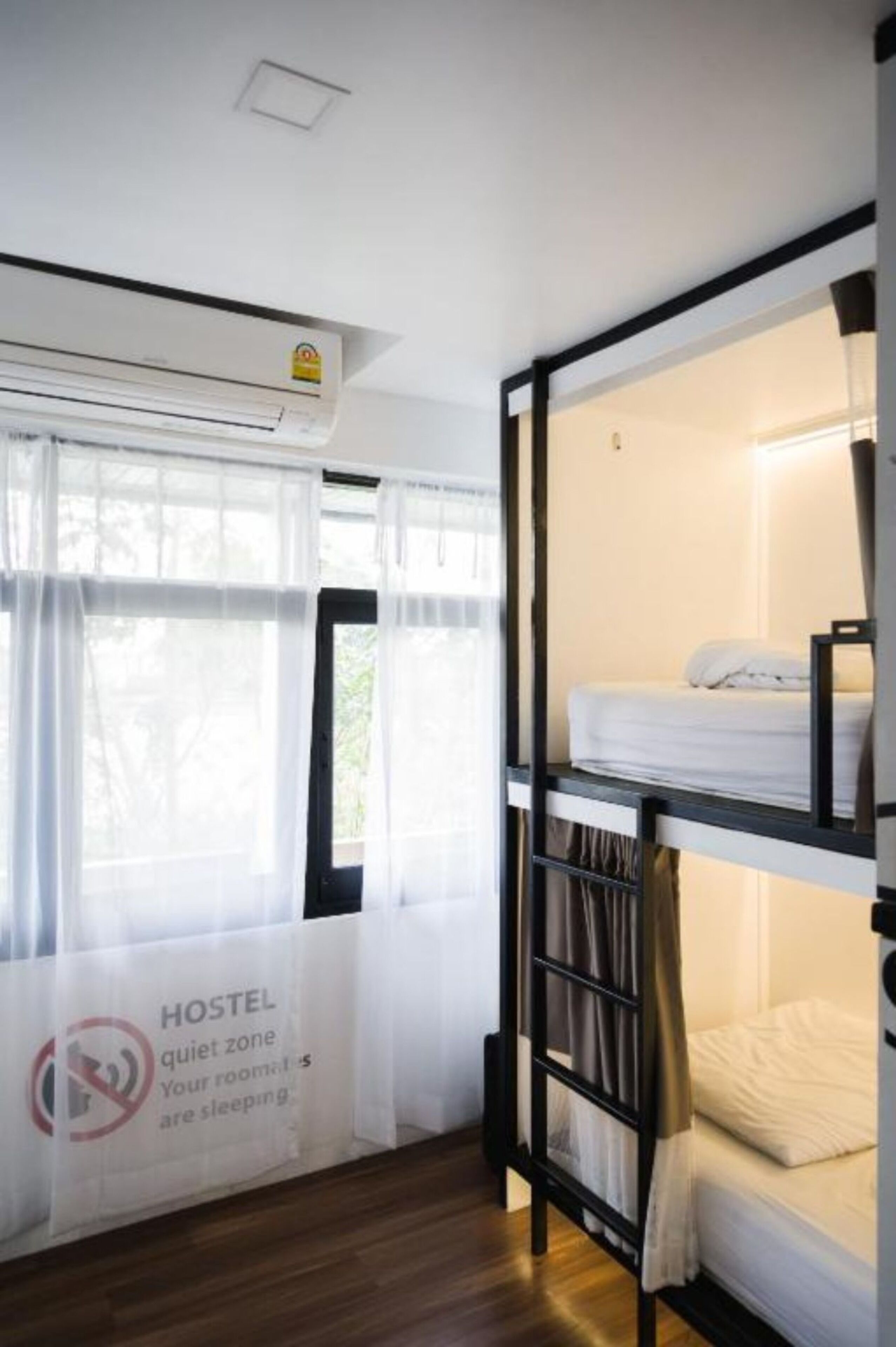 A hostel room with bunk beds, a window with sheer curtains, and a sign indicating a quiet zone.