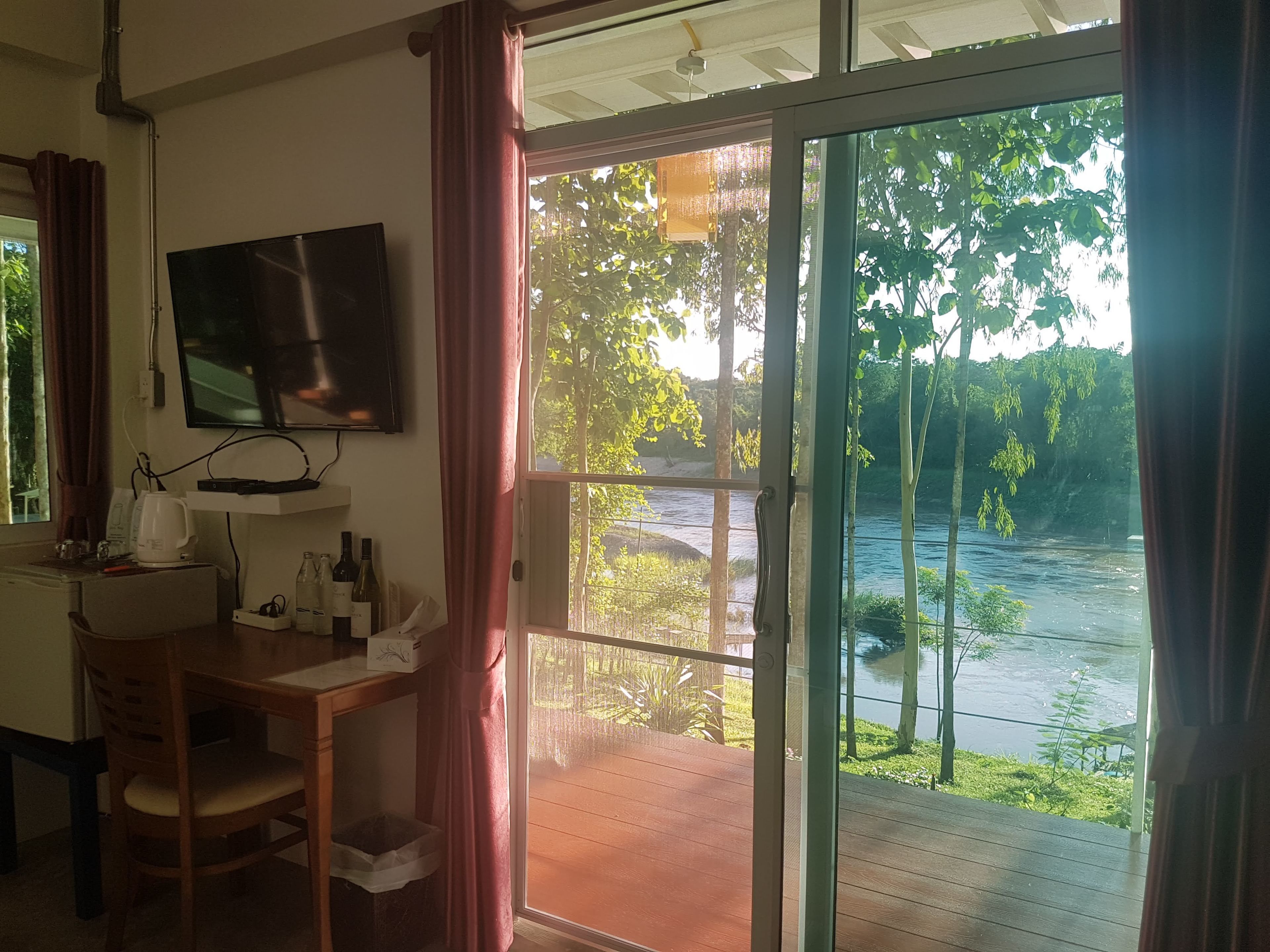 A room with a TV mounted on the wall, a wooden desk with a chair, a small table with bottles, and a view of a river through a glass door.