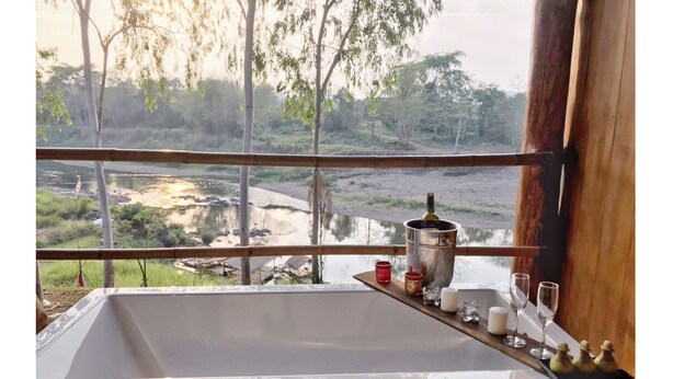 A bathroom with a large sink, a wooden shelf with candles and glasses, and a view of a river and trees. - Nan De Panna Riverside Homestay (Phu Phiang)