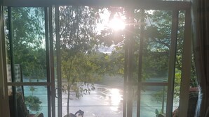 A view through a window with a wooden deck, a metal railing, and a view of a body of water with trees and a structure in the distance. - Nan De Panna Riverside Homestay (Phu Phiang)