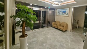 Un vestíbulo moderno con un gran árbol en maceta, una entrada con puerta de cristal, dos sofás beige y un cuadro con una vista urbana enmarcada en la pared.