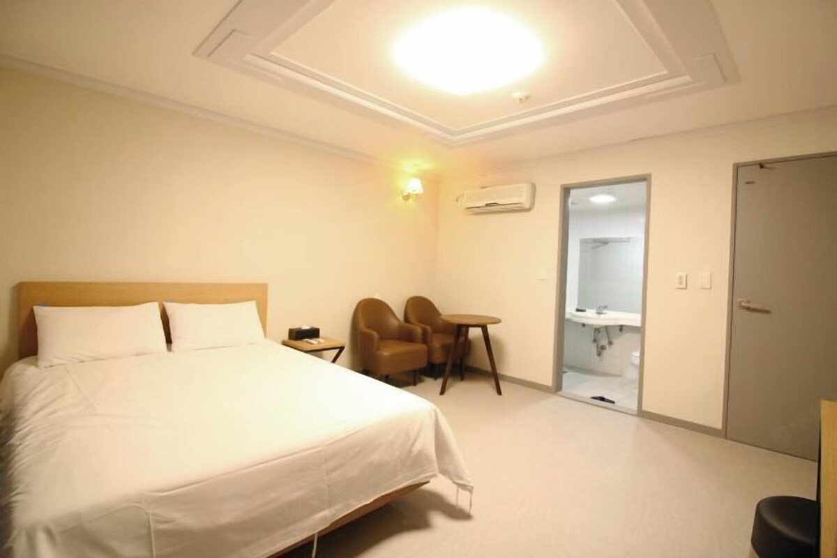 A hotel room with a bed, a desk, two chairs, a small table, a bathroom with a sink and mirror, and a door.