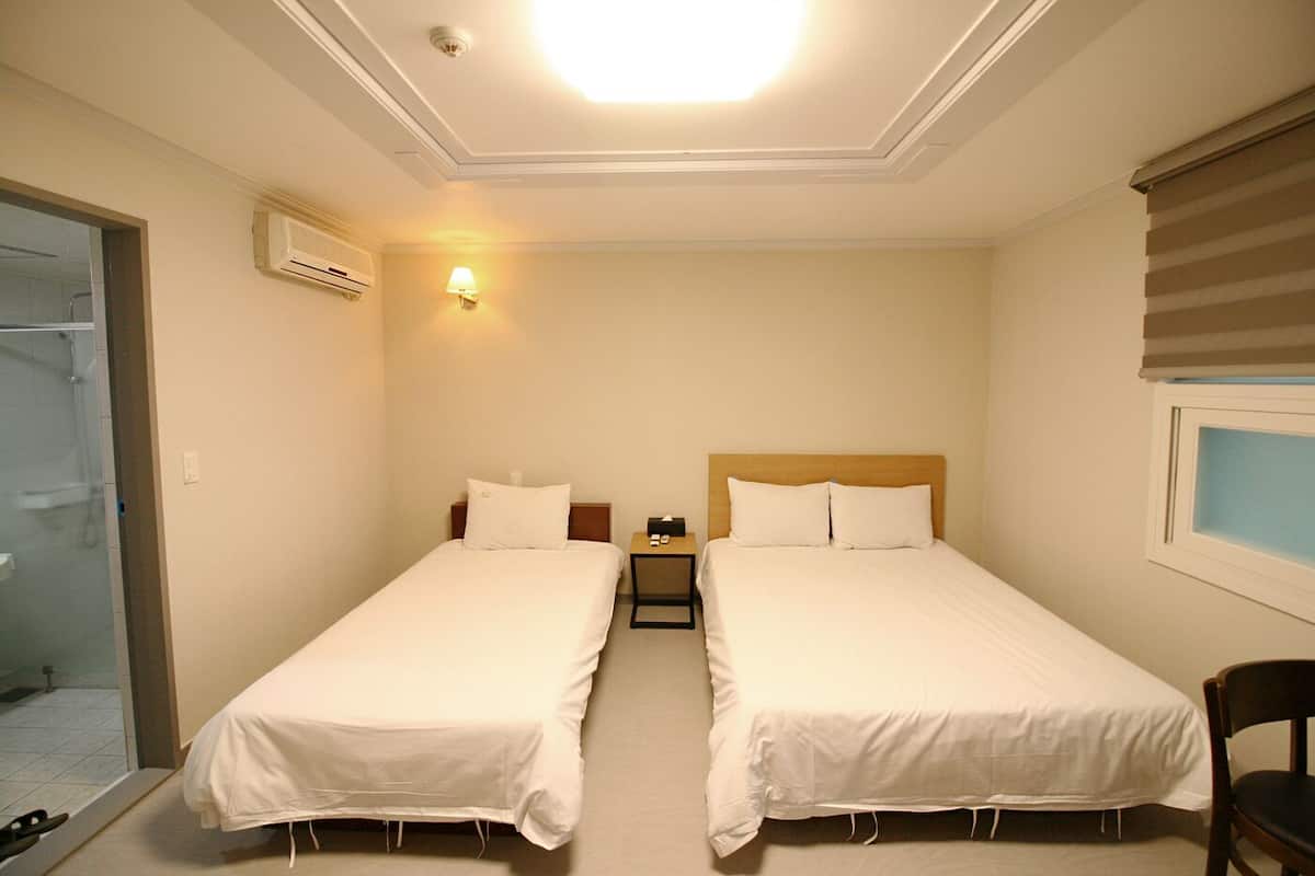 A hotel room with two single beds, a nightstand, a lamp, and a window with blinds.