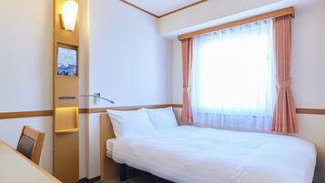Camera d'albergo con un letto, una scrivania, una sedia, una lampada a parete e una finestra con tende.