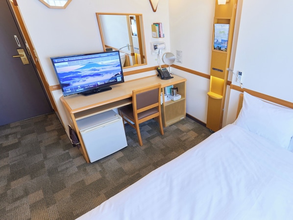 A hotel room with a bed, a desk with a computer, a small refrigerator, a bedside table with a lamp, and a mirror.