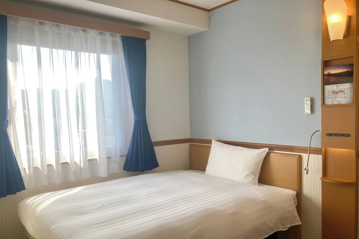 Ein Hotelzimmer mit einem Bett, Vorhängen, einer Wandleuchte und einer kleinen Ablagefläche mit einem Bild.