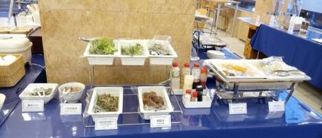 Uma mesa de buffet com diversos alimentos e acompanhamentos, cercada por mesas e cadeiras.