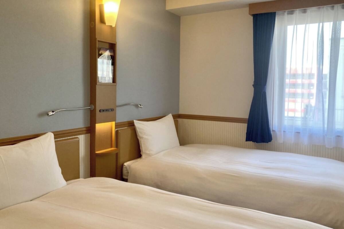 Ein Hotelzimmer mit zwei Betten, einem Nachttisch, einer Wandlampe und einem Fenster mit Vorhängen.