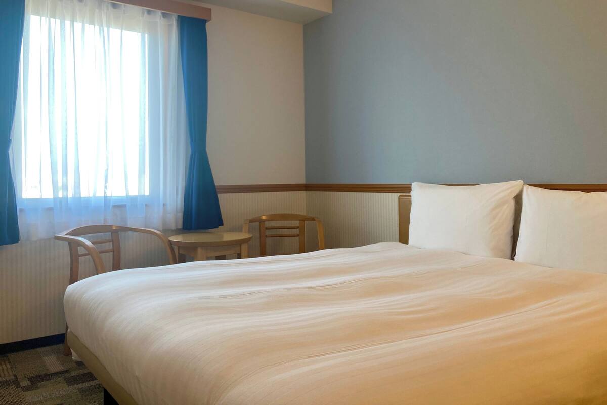 Ein Hotelzimmer mit Bett, einem kleinen Tisch, einem Stuhl und einem Fenster mit Vorhängen.