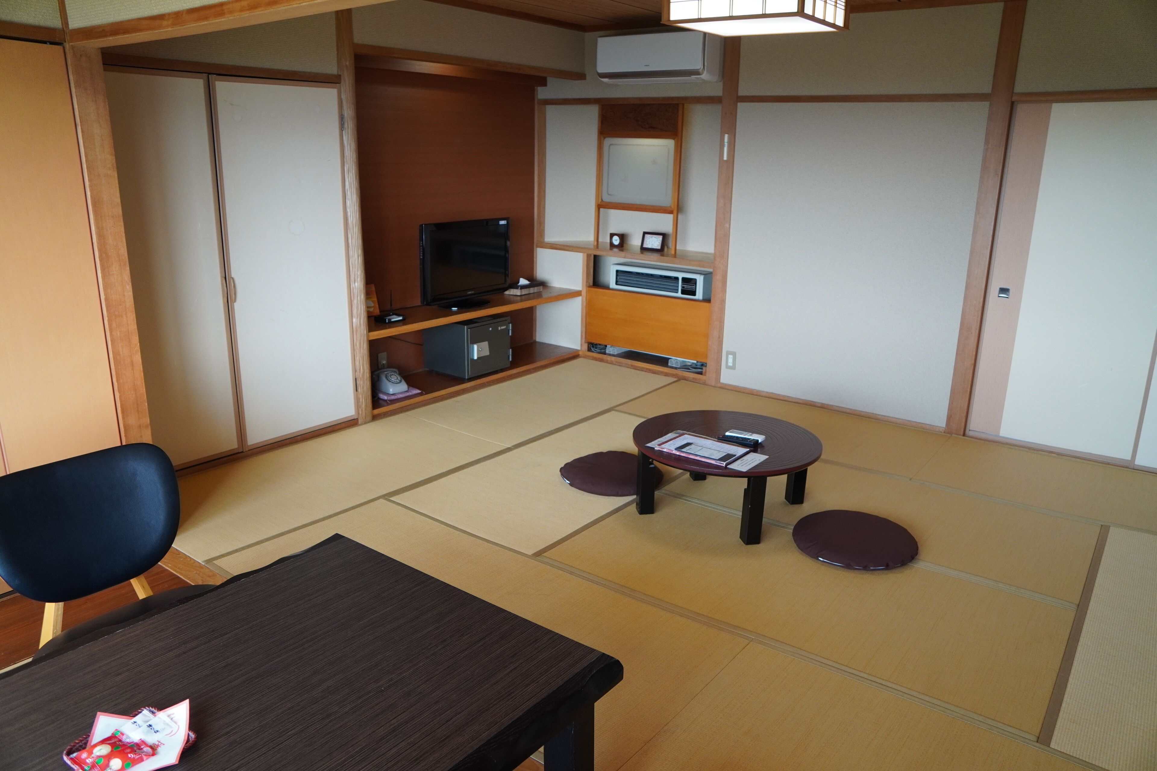 A traditional Japanese room with tatami flooring, a low table, a black chair, and a television on a shelf.