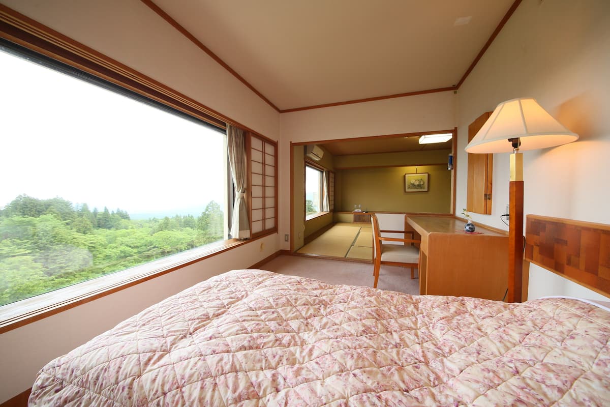 Une chambre d’hôtel avec un grand lit, un bureau, une chaise et une vue sur une forêt, visible par la fenêtre.
