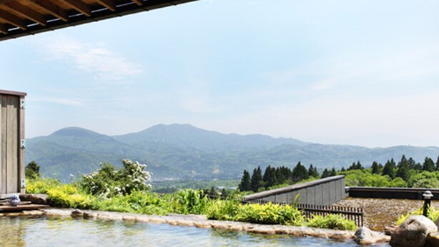 Une source thermale de pierres, dotée d’une terrasse en bois, entourée de verdure et avec des montagnes en toile de fond.