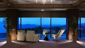 Une pièce spacieuse avec de grandes fenêtres offrant une vue sur les montagnes et un lac au crépuscule. On y trouve deux fauteuils en cuir et une petite table sur laquelle est posé un vase de fleurs.