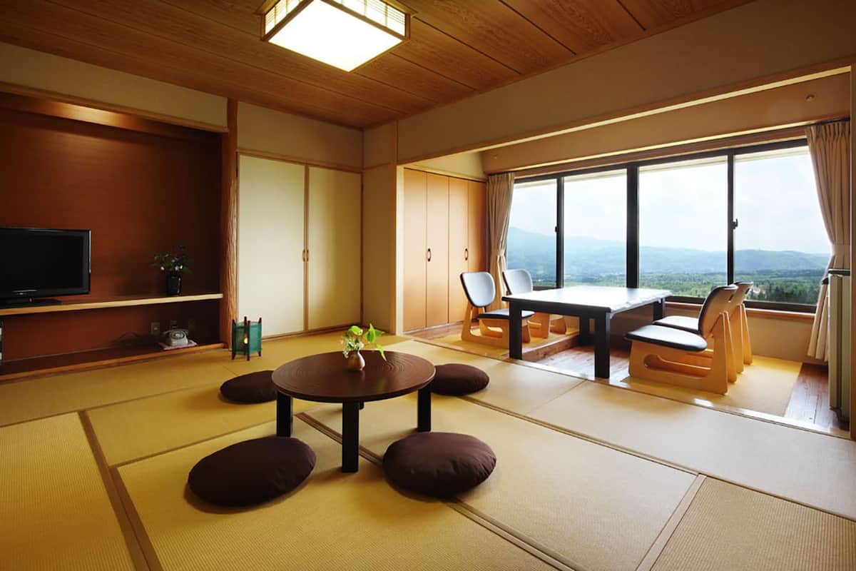 Une pièce de style japonais traditionnel, avec un sol recouvert de tatamis, une petite table basse avec des coussins, un téléviseur et une vue sur les montagnes.
