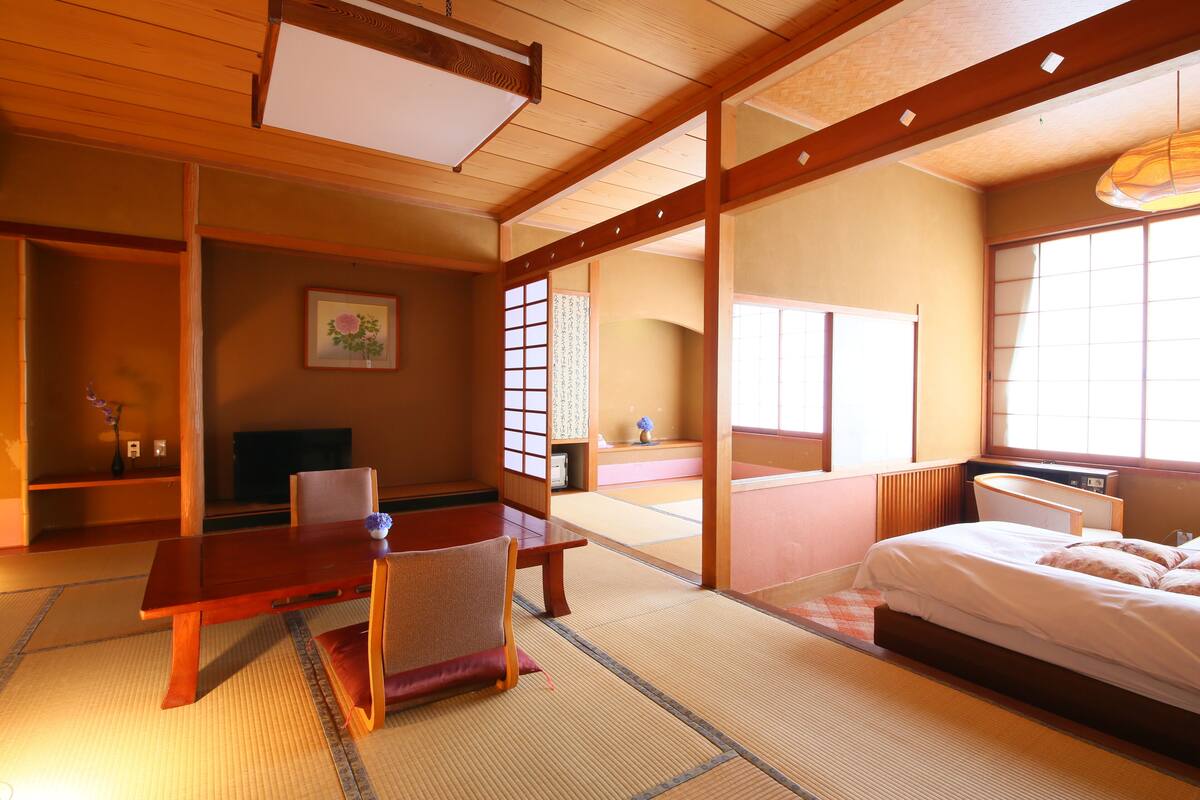 Une pièce de style japonais traditionnel, avec un sol recouvert de tatamis, une petite table en bois, un lit avec une literie blanche et des portes coulissantes.