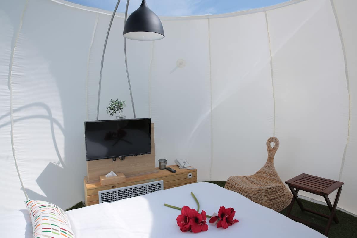 A tent-style room with a bed, a TV, a small table, a chair, and a hanging lamp.