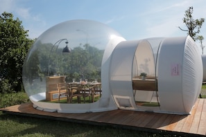 Una estructura abovedada transparente con una mesa y sillas de madera en su interior, rodeada por una terraza de madera.