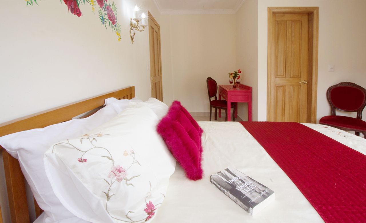 A neatly made bed with a floral bedspread, a red throw blanket, and a magazine on the bed. There is a wooden headboard, a red chair, and a wooden door in the background.