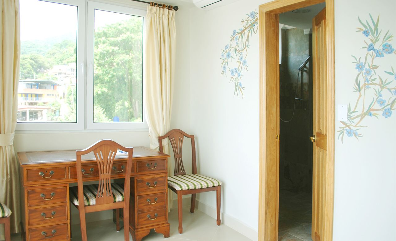 Una habitación con un escritorio de madera, dos sillas, un ventanal con cortinas y una puerta que da a una zona de ducha.