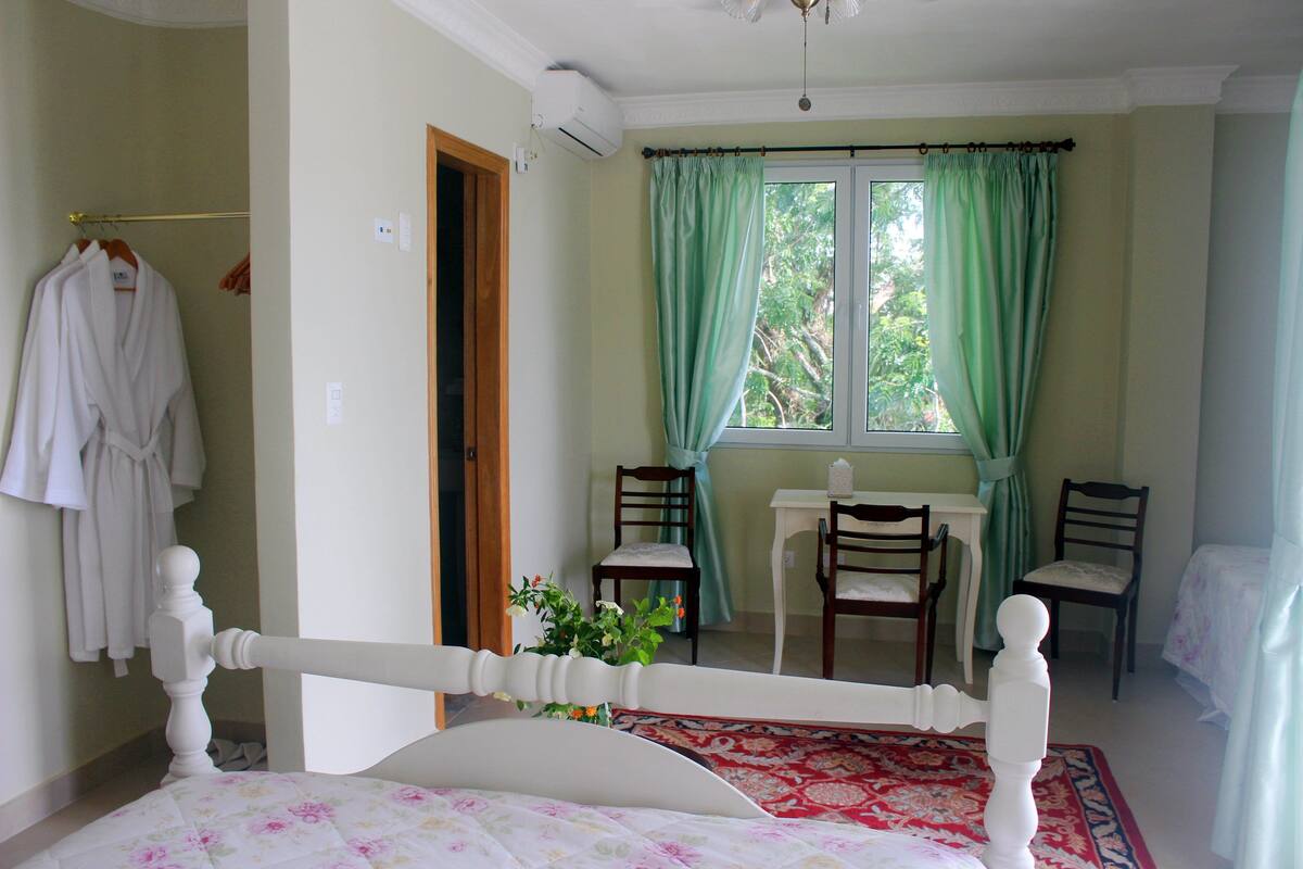 A bedroom with a bed, a desk, two chairs, a window with curtains, and a hanging robe.