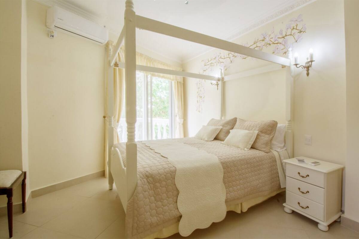 A four-poster bed with a quilted bedspread, a bedside table with drawers, and a wall-mounted light fixture.