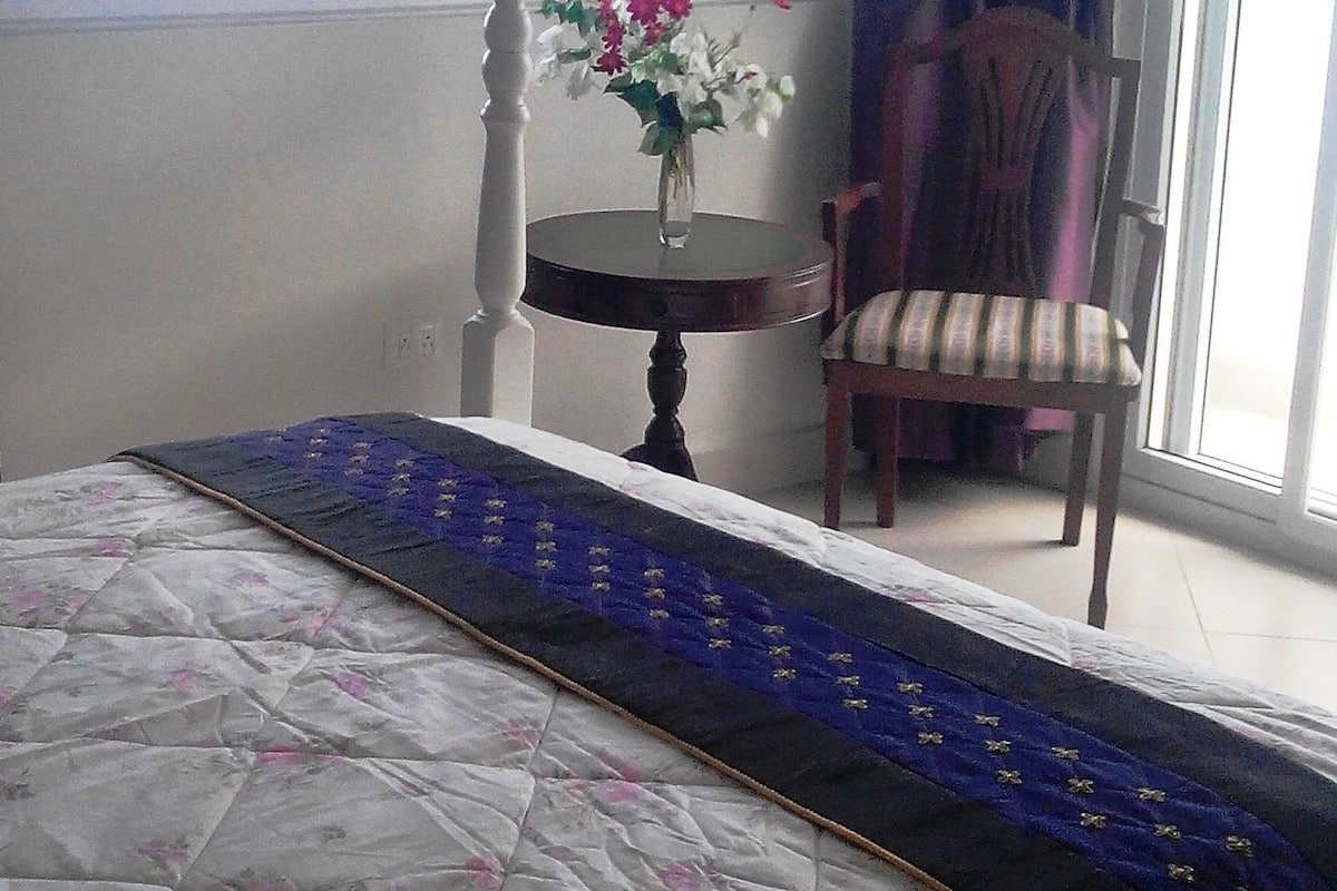 A bedroom with a bed, a chair, a small table with a vase of flowers, and a window with curtains.