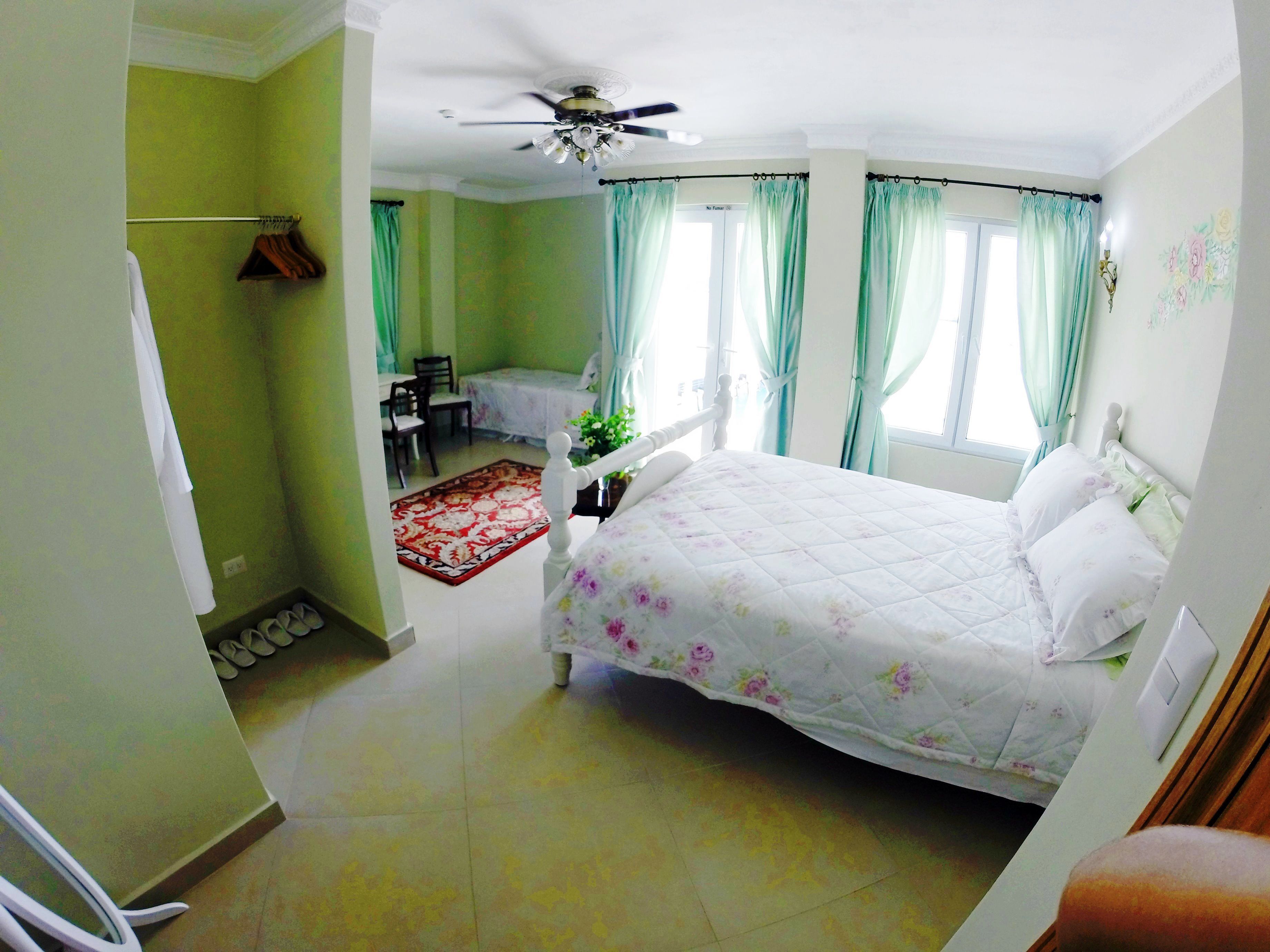 Une chambre à coucher avec un lit, un ventilateur de plafond, une fenêtre avec des rideaux et un petit bureau avec une chaise.