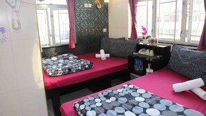 Habitación de hotel con dos camas, cada una con cubrecama rojo y almohadas estampadas. Hay una mesita con lámpara, teléfono y un florero con flores. La habitación tiene ventana con cortinas y vistas a edificios.