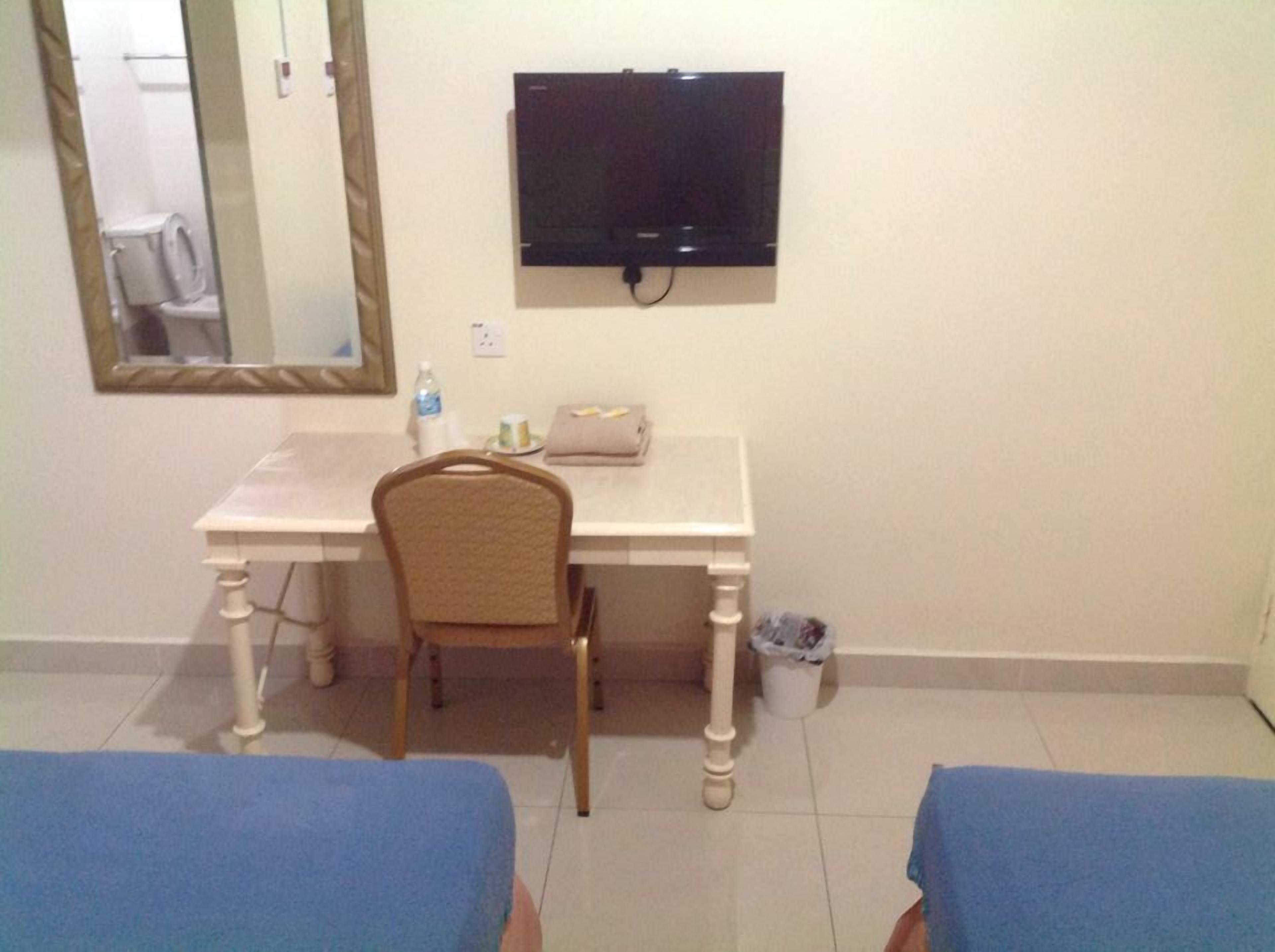 A room with a white desk, a chair, a television mounted on the wall, a mirror, and two beds with blue covers.