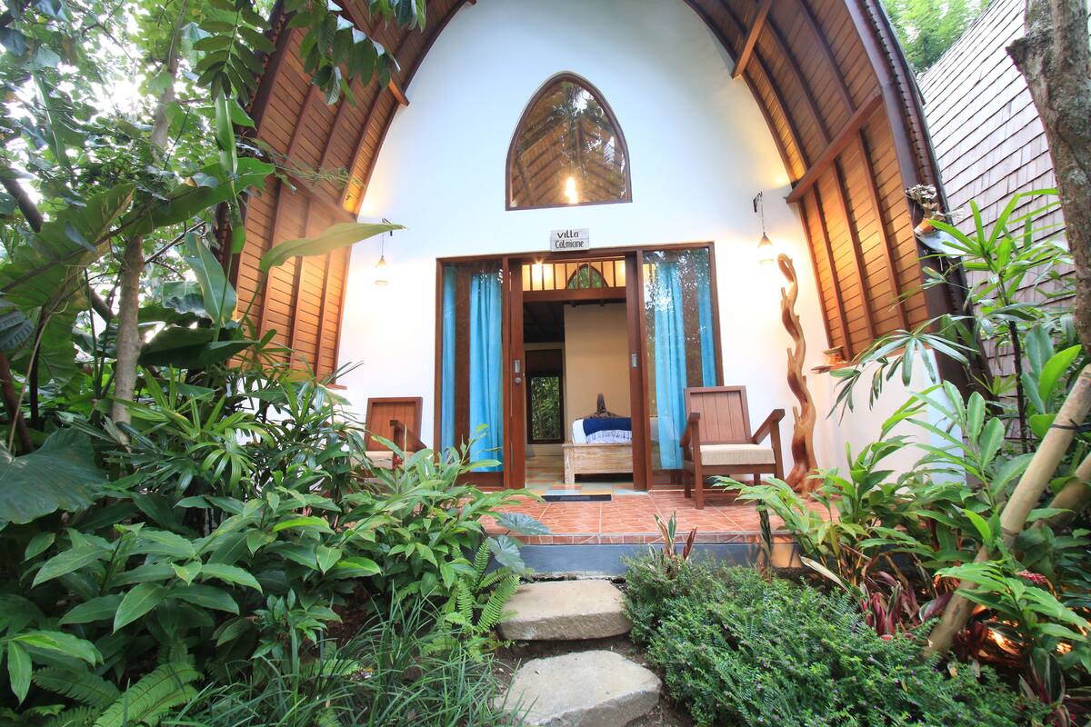 A villa with a wooden exterior, a stone staircase leading to a wooden door, and a sign above the door reading "Villa Calimene".