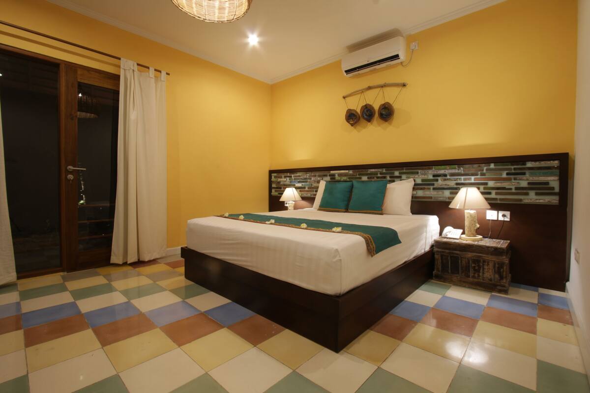 A hotel room with a checkered floor, a bed with a wooden headboard, a nightstand with a lamp, and a sliding glass door.