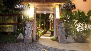A pathway leading to a thatched-roof structure with two umbrellas, surrounded by tropical plants and stone statues. - Green Bird Villa (Ubud)