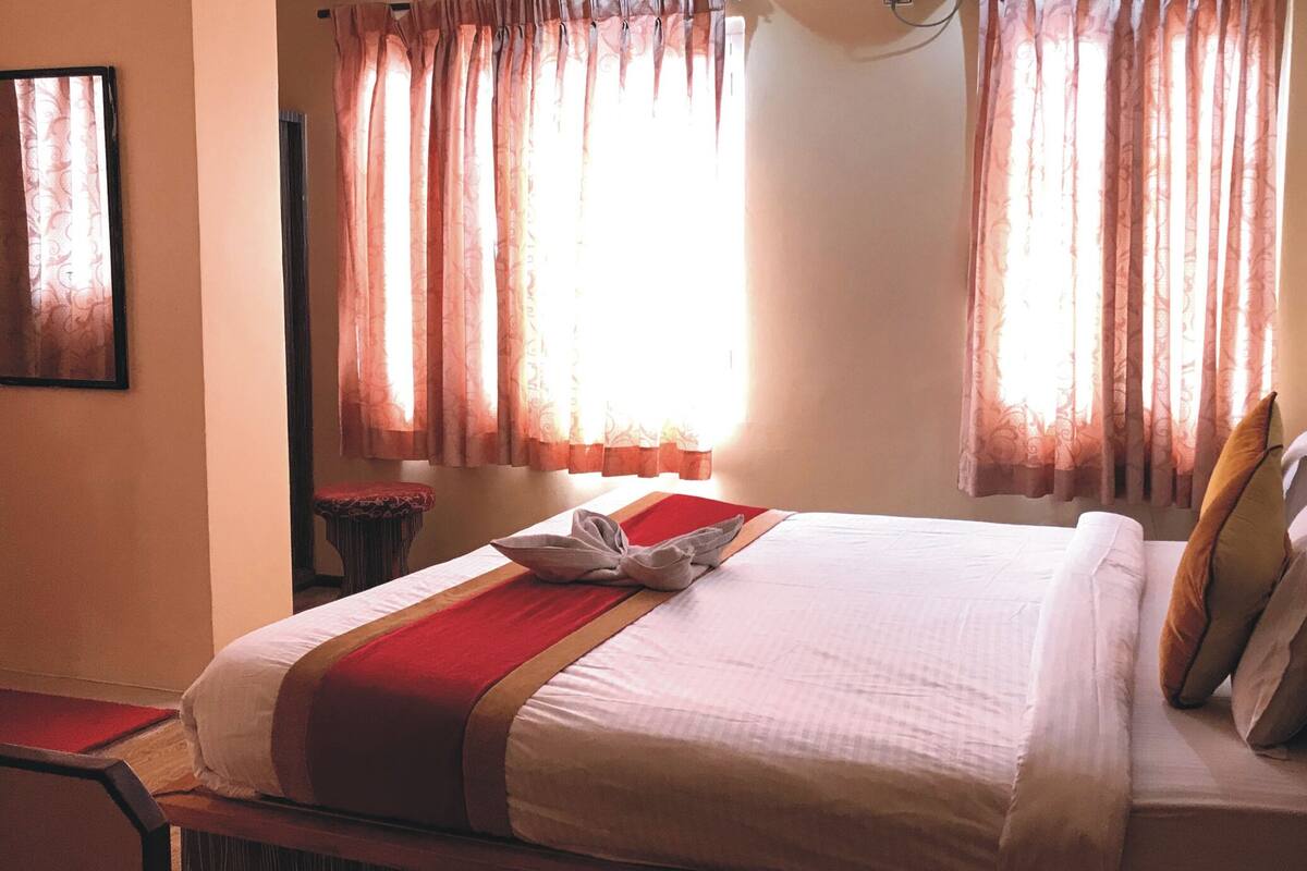 A neatly made bed with a red and white bedspread, a pillow, and a folded towel. There are two windows with sheer curtains, a bedside table with a vase, and a chair.