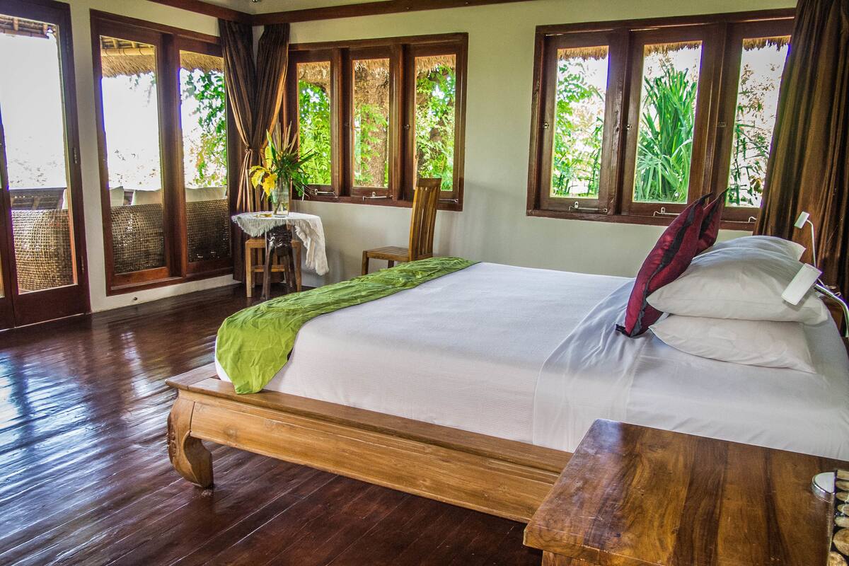 A spacious room with a large bed, wooden flooring, and large windows with curtains.
