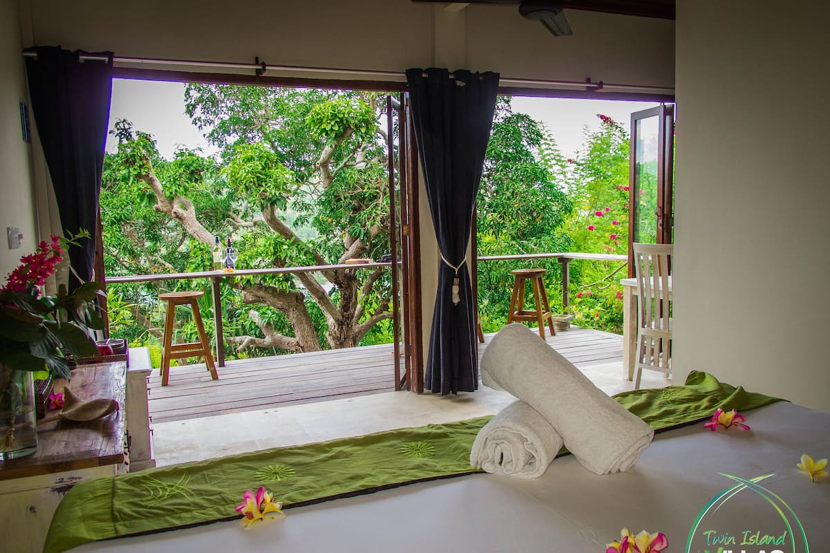 A room with a bed, a view of a balcony with a wooden railing, and a large tree outside.