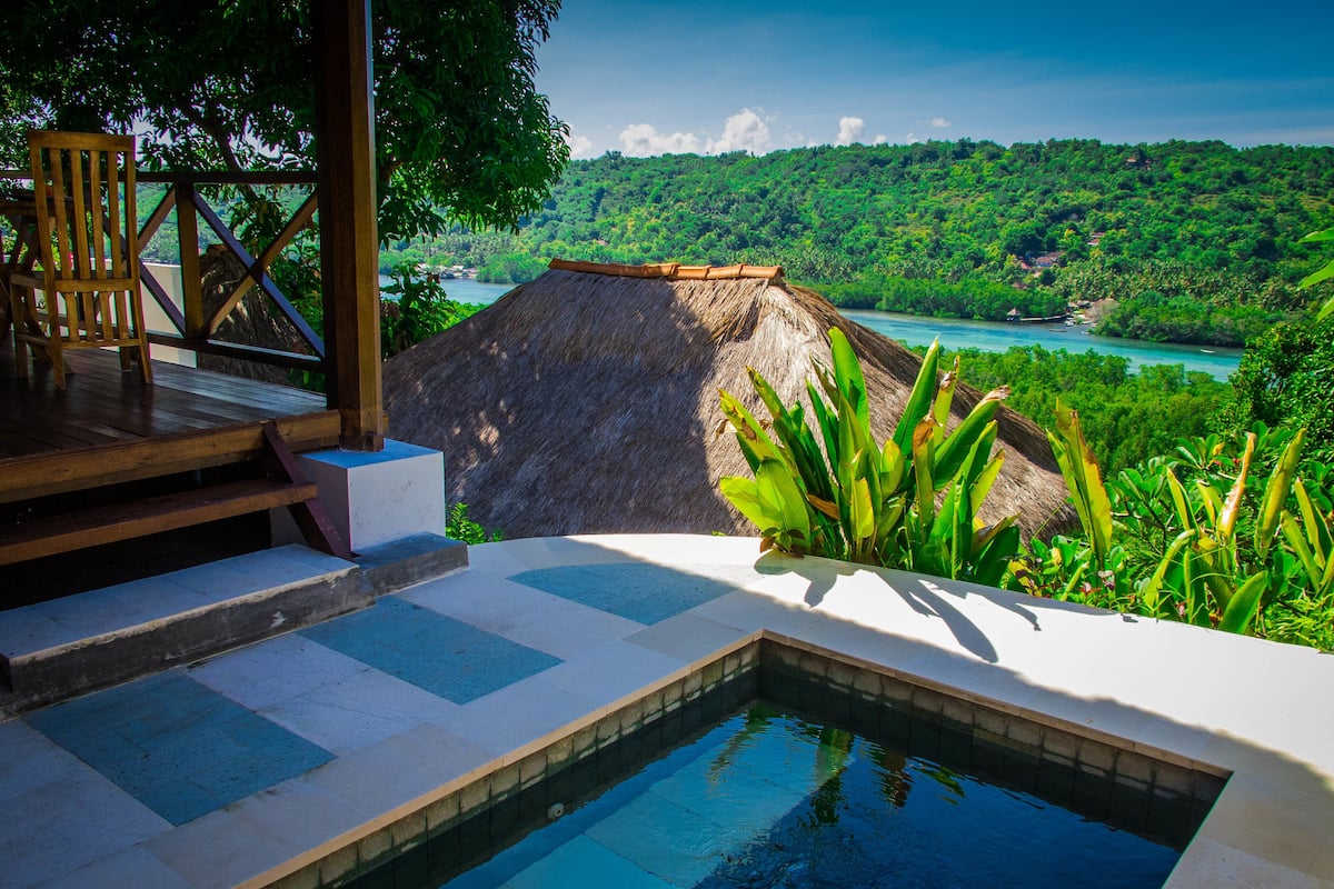 A pool area with a view of a tropical landscape, a wooden deck, and a thatched roof structure.