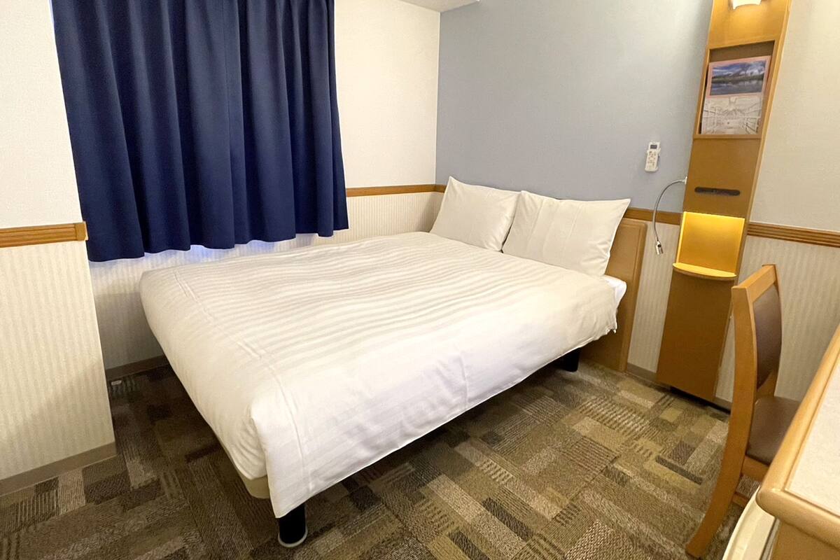 A hotel room with a bed, a desk, a chair, a nightstand, and a window with curtains.