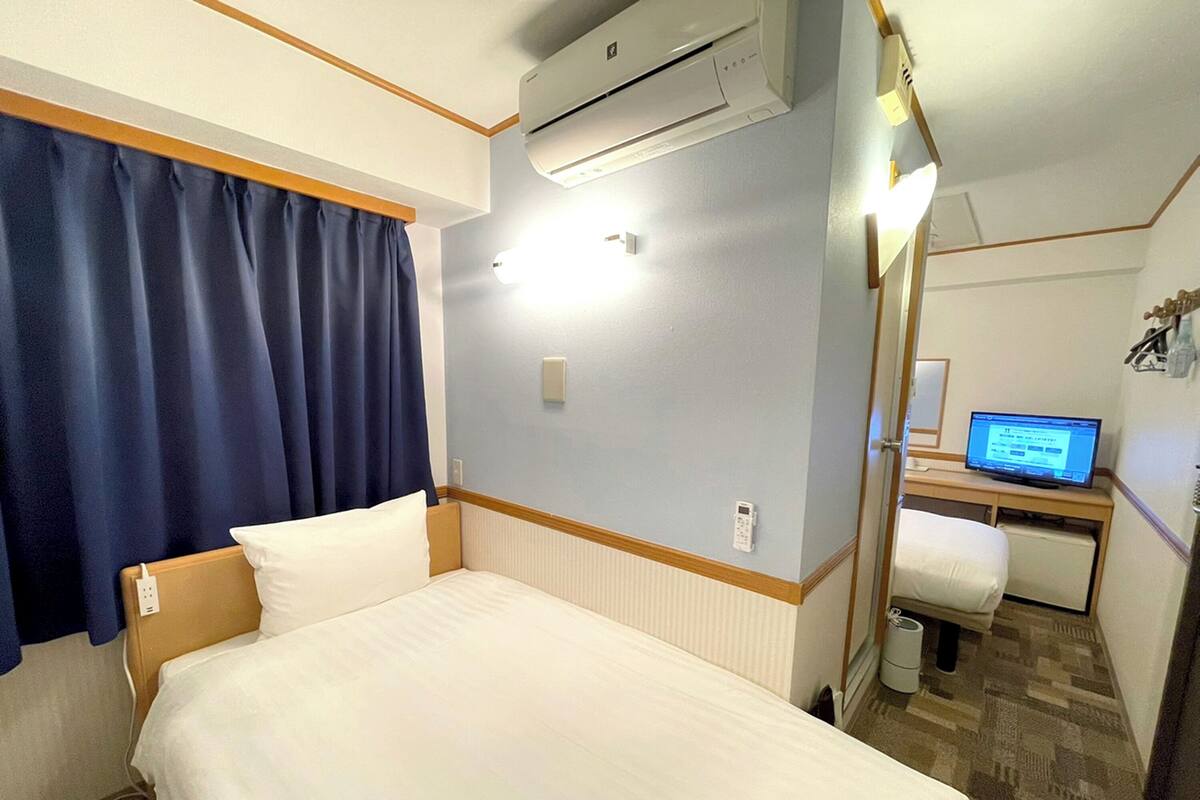 A hotel room with a bed, a desk with a computer, an air conditioning unit, and a window with curtains.