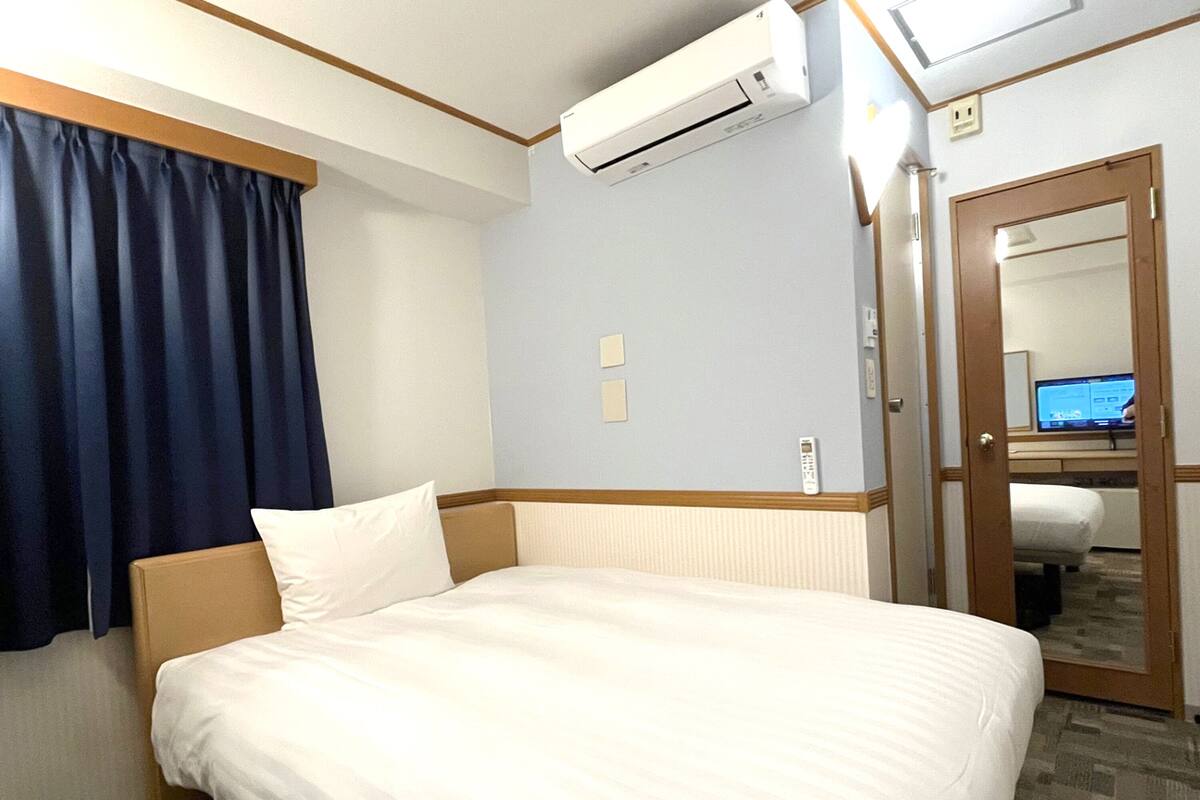 A hotel room with a bed, an air conditioning unit, a desk with a computer, and a door leading to another room.