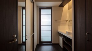 Un pasillo estrecho con una puerta de madera oscura a la izquierda, una puerta de vidrio esmerilado al final y un baño blanco con espejo y lavamanos a la derecha.