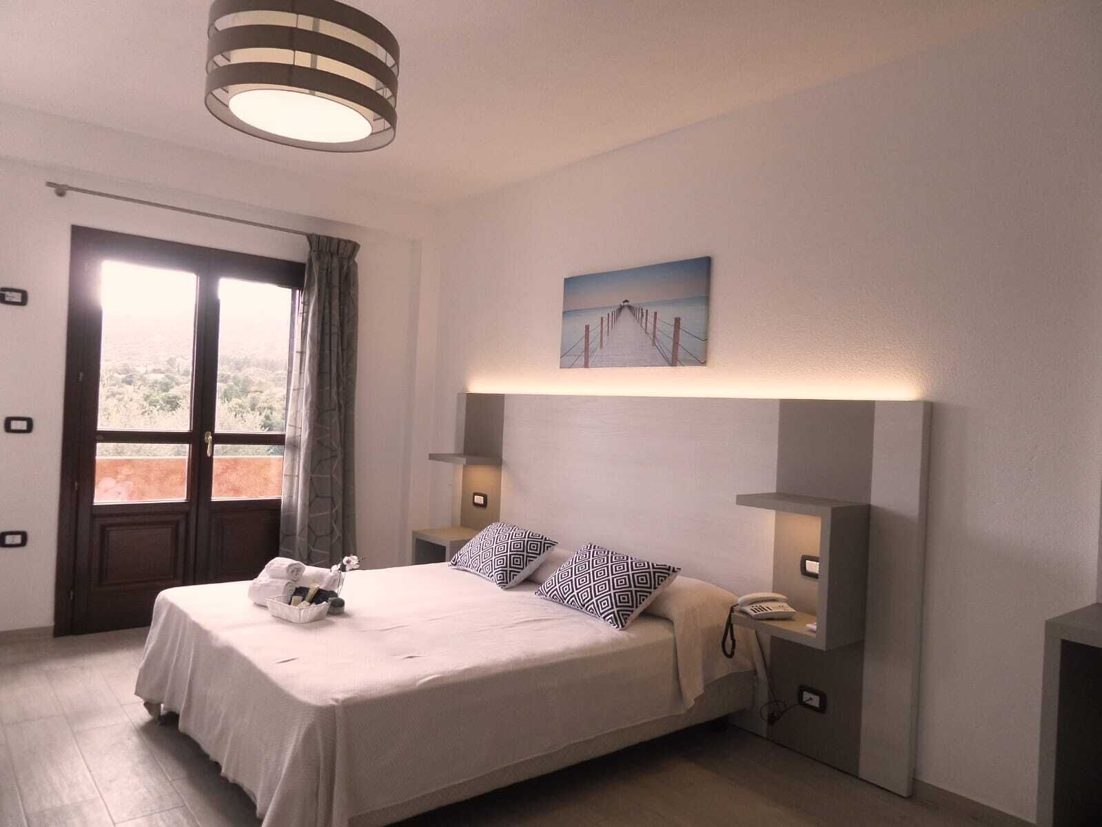 A neatly arranged bedroom with a bed, bedside tables, a painting, and a view through the window.