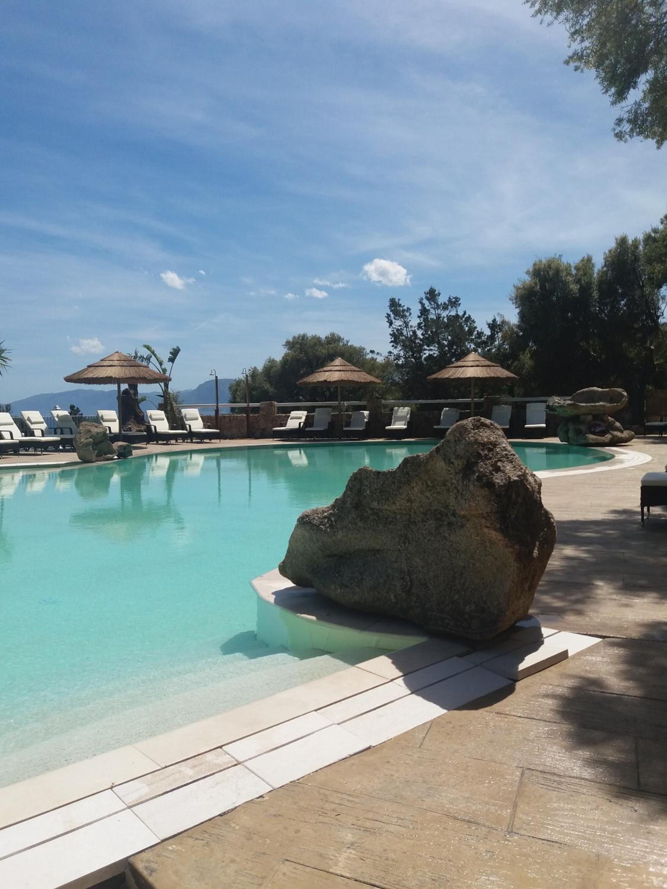 A clear pool with lounge chairs and umbrellas, surrounded by trees and a view of the ocean.