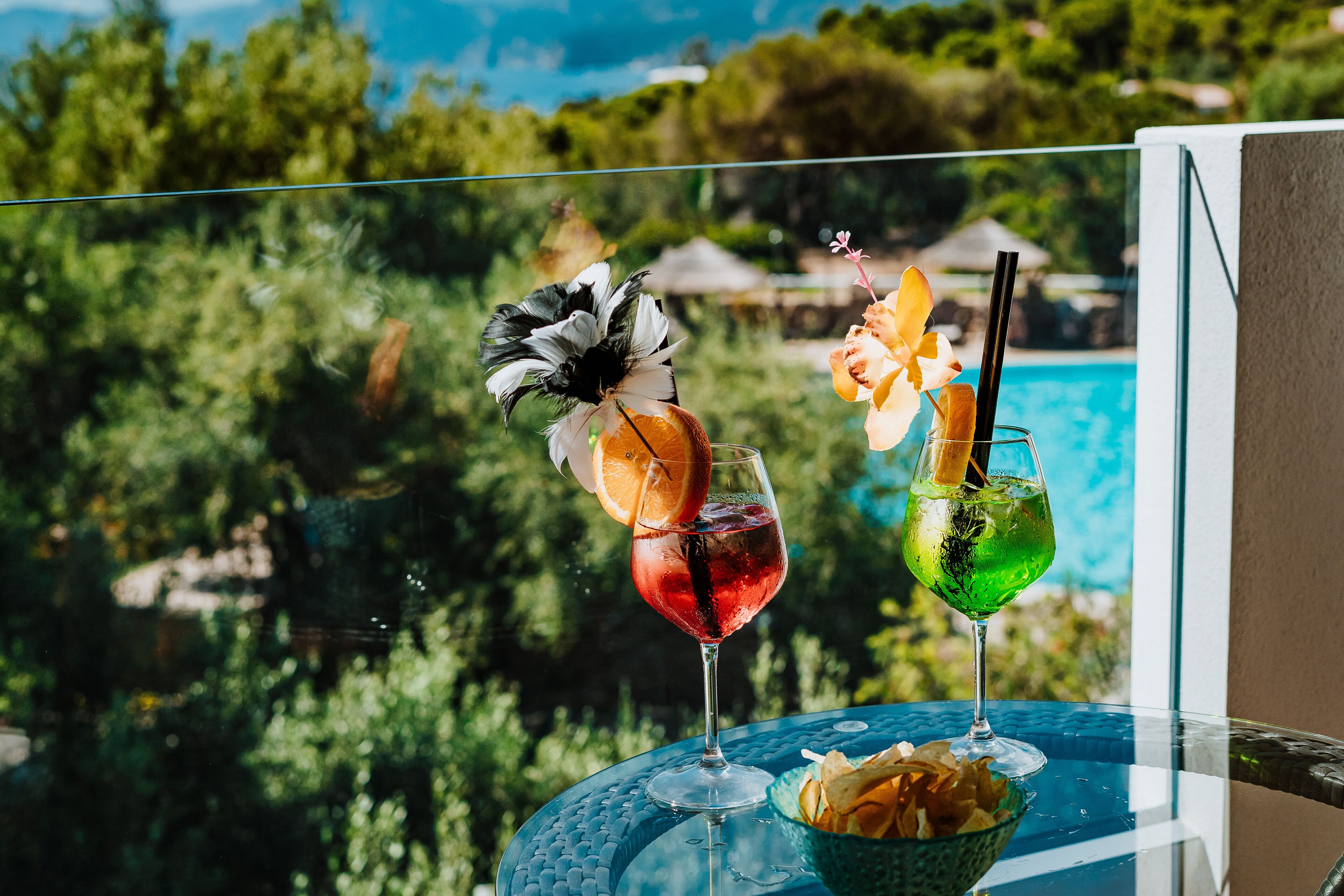 Two drinks on a glass table with a pool view.