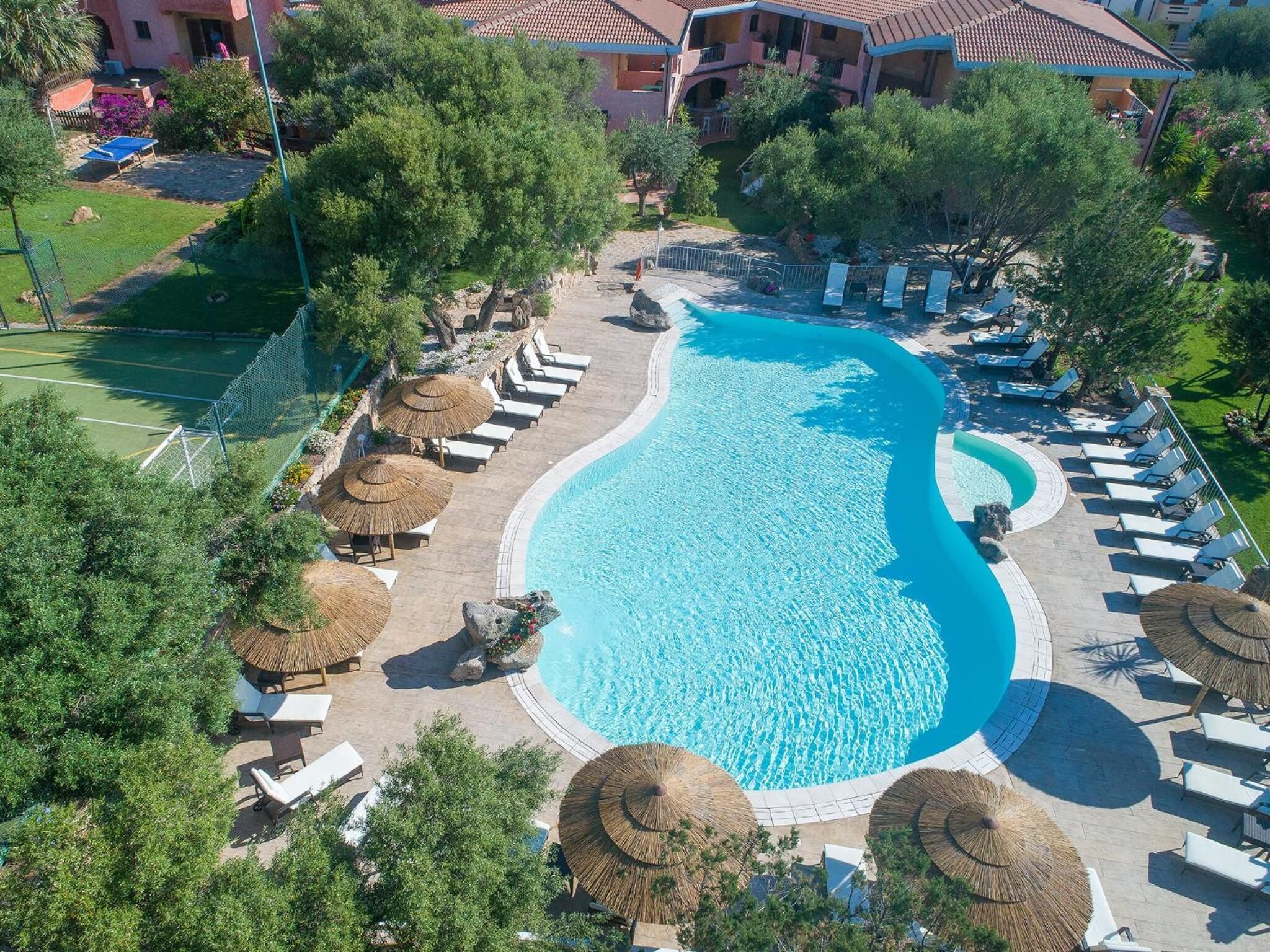 A large, curved swimming pool surrounded by lounge chairs and thatched umbrellas, with a tennis court and greenery in the background.